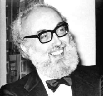 Monochrome portrait of a smiling man with a full white beard and glasses wearing a dark jacket and bow tie against a blurred background