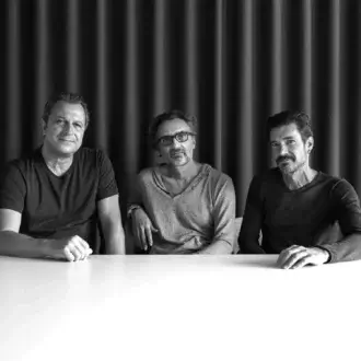 Three men stand behind a light horizontal surface with hands resting on top against a dark ribbed backdrop