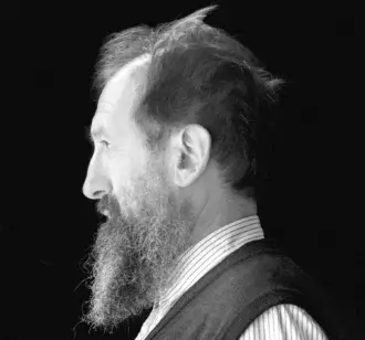 Monochrome portrait of a man with a beard facing left, wearing a striped collared shirt and dark vest against a dark background