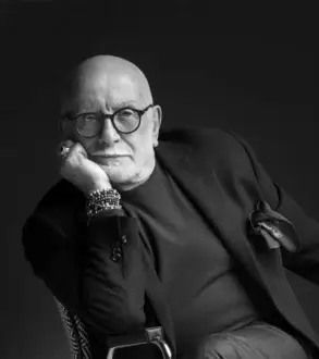 Man in charcoal blazer seated in chair, wearing glasses, ring, bracelets, against dark backdrop, portrait with focused lighting.