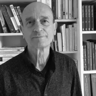 Grayscale portrait of a man between bookshelves, displaying book titles including “Hard Times” and Italian literature, wearing a dark textured shirt.