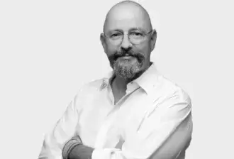 Black and white portrait of Klaus Nolting, a man with a gray beard and glasses, wearing a button-down shirt and woven bracelets.