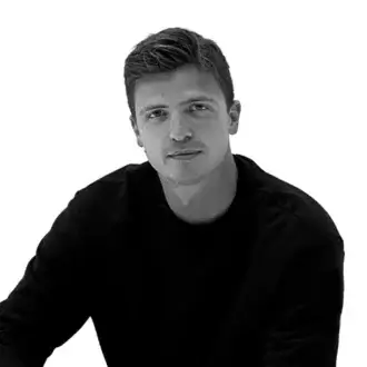 Black and white portrait of Marco Oggian, wearing a dark crew neck shirt, with textured hair and a subtle expression against a white background.