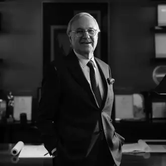 Pierre Yves Rochon portrait, silver hair, dark suit, floral lapel pin, standing in office with desk, drawings, artwork, soft lighting.