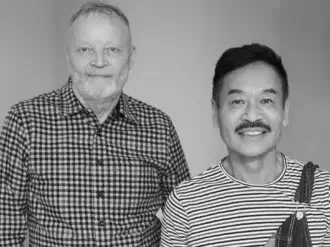 Black and white portrait of two designers, Yabu Pushelberg, with checkered and striped shirts against a gray backdrop.