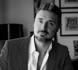 Black and white portrait of Massimiliano Raggi in a patterned black blazer and white shirt, with framed art in background.