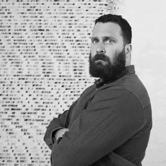 Man with beard, brown shirt, against dense white text backdrop, evenly lit, creating contrast between figure and informational pattern.
