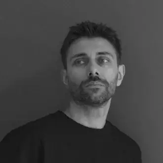 Man portrait, grayscale, short dark hair, black sweatshirt, neutral expression, light gray eyes, solid gray background.