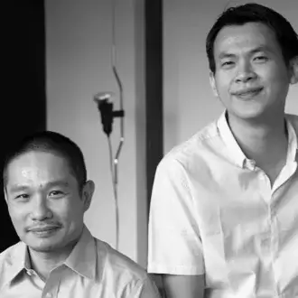 Black and white portrait of Voon Wong & Benson Saw, both wearing light shirts, against a minimalist interior with a suspended microphone.