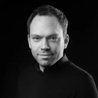 Black and white portrait of Daniel Becker, dark hair, fair complexion, wearing a black crew-neck sweater against a black background.
