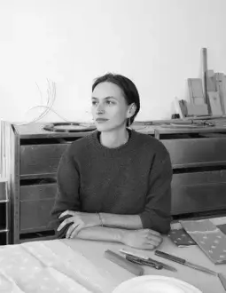 Black and white portrait of Julie Richoz seated at a table with tools, a patterned fabric, and a metal cabinet backdrop.