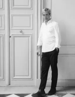 Black and white portrait of Sam Baron in white shirt, standing before paneled door with rectangular sections and tiled floor.
