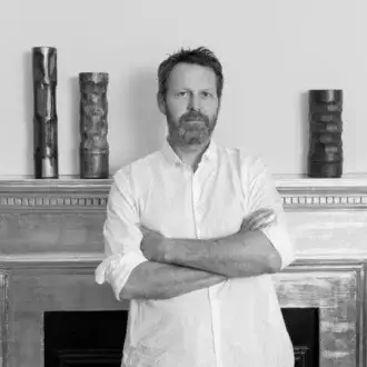 Martino Gamper portrait with textured cylindrical sculptures on a weathered mantel, soft lighting, and neutral wall backdrop.