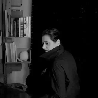 India Mahdavi portrait, grayscale, seated at desk with books on metal shelving, wearing dark collared shirt, soft directional lighting.