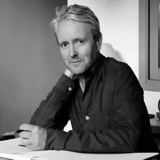 Ronan Bouroullec portrait, black and white photograph, seated man with light blonde hair at a white desk with pen and notebook.