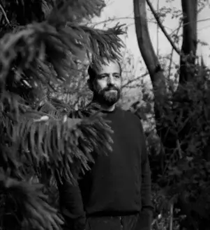 Black and white portrait of Michael Anastassiades in a dark sweater, framed by evergreen foliage, with a serious expression and diffused natural lighting.