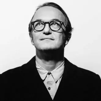 Black and white portrait of Massimiliano Locatelli, a man in a blazer and glasses looking up with a thoughtful expression against a white background.