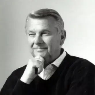 Black and white portrait of Tito Agnoli, a man with gray hair wearing a dark sweater and striped collared shirt, hand resting on chin.