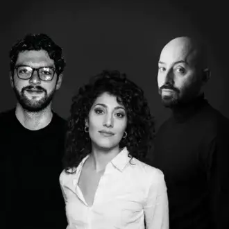 Black and white portrait of three people—man, woman, and man—with curly hair, glasses, and beards against a dark gray backdrop.