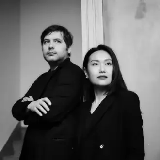 Black and white portrait of two partners in black blazers, standing before a distressed plaster wall with a visible staircase.