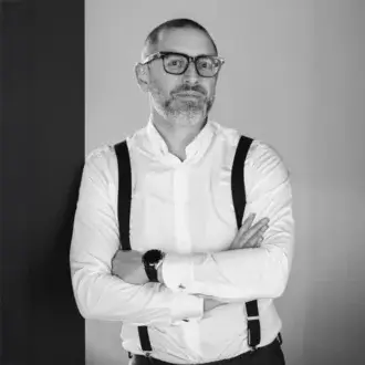 Black and white portrait of Andrea Citterio, a man with gray beard wearing white shirt, dark suspenders, and framed glasses, arms crossed.