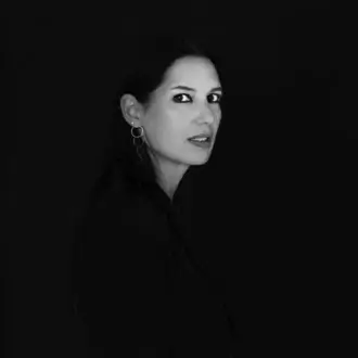 Black and white portrait of Elisa Ossino, three-quarter profile, dark collared shirt, silver hoop earrings, minimalist composition, diffused lighting.