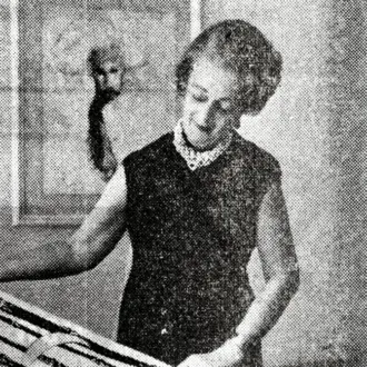 Woman behind table with rectangular objects, wearing dark dress and beaded necklace, gesturing in diffused light interior.