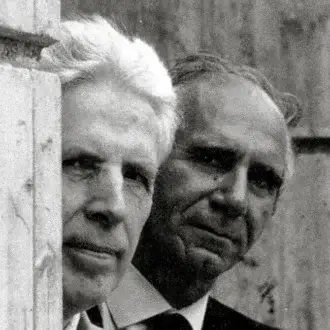 Black and white portrait of two men in suits, framed by a weathered wood post, with high contrast and blurred outdoor background.