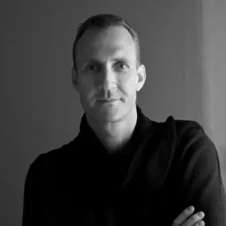 Black and white portrait of Michael Geldmacher, a man with short hair, wearing a dark knit hooded sweater, arms crossed, against a light gray backdrop.