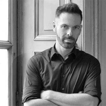 Black and white portrait of David Obendorfer, a man with short dark hair and beard, wearing a dark button-down shirt indoors by a window.