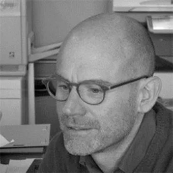 Bald man Emanuele Zenere in glasses and dark shirt, neutral expression, office background with boxes, printer, monitor, diffused lighting.
