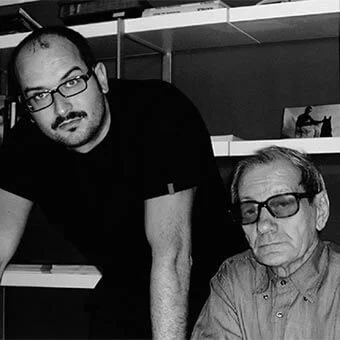 Black and white portrait of two men indoors, featuring a man in glasses with a white surface, and an older man with white hair, bookshelves visible.