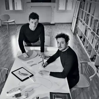 Two people flank a light surface displaying sketches, rectangles, a tablet image and papers in a room with wood floors