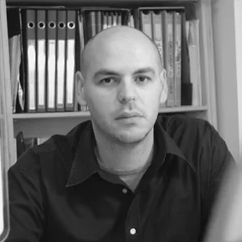 Grayscale portrait of a person with shaved head and stubble wearing a dark collared shirt before a bookshelf filled with dark files