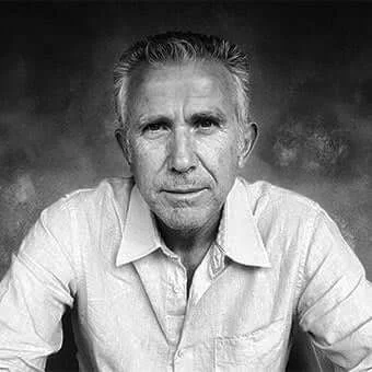 Black and white portrait of Aldo Greco, a man with gray hair, wearing a light linen shirt, direct gaze, blurred dark gray background.