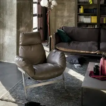 Leather lounge chair, brushed silver frame, high back, rounded arms, gray & blue rug, gray sofa, wood bookcase, concrete wall.