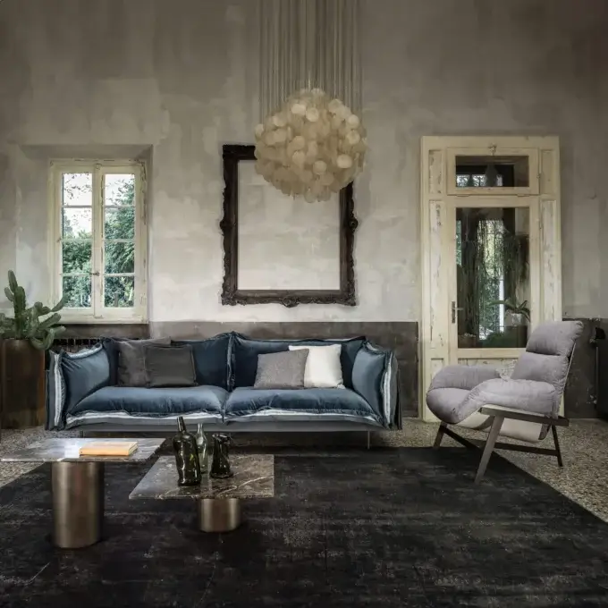Blue velvet sofa with oversized cushions, dark gray rug, brass and marble coffee tables, ornate mirror, and chandelier in a living room.