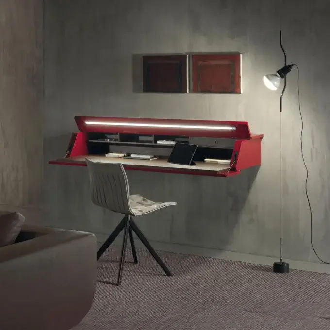 Fold-down desk, red exterior, light wood work surface, integrated LED light, mounted on gray textured wall with artworks, modern chair nearby.