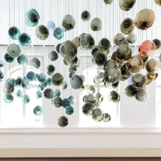 Bocci lighting installation, hand-blown glass orbs, varying teal, green, gray, amber, and pink hues, suspended in a dense, cloud-like arrangement.