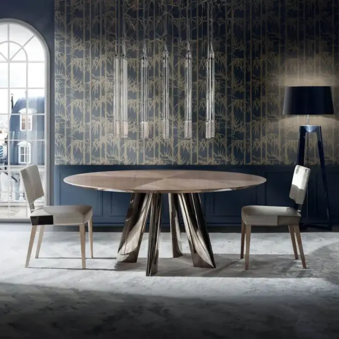 Round dining table with walnut top and metal base, paired with beige upholstered chairs, on a gray rug in a room with blue bamboo wallpaper.