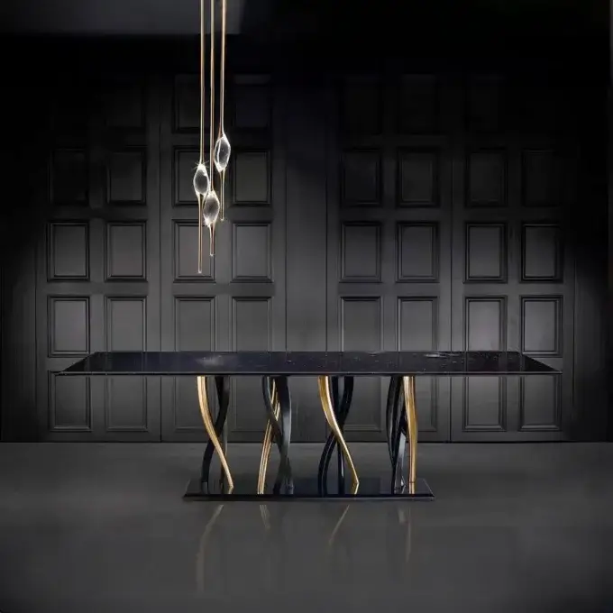 Dining table, black marble, with three twisted gold metal legs, under a multi-tiered glass chandelier, against dark gray paneled walls.