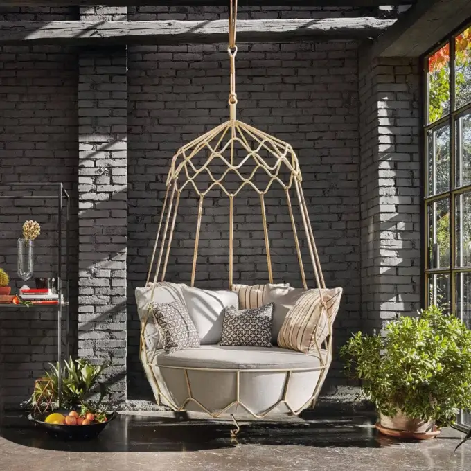 Rattan chair, light beige cushion, geometric gold frame hangs in room with gray brick walls, plants, and natural light.