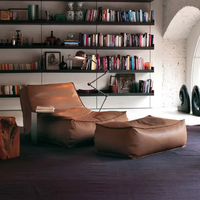 Leather armchair verzelloni, brown, low-slung with ottoman on purple rug, brass floor lamp, gray shelves with books and decor.