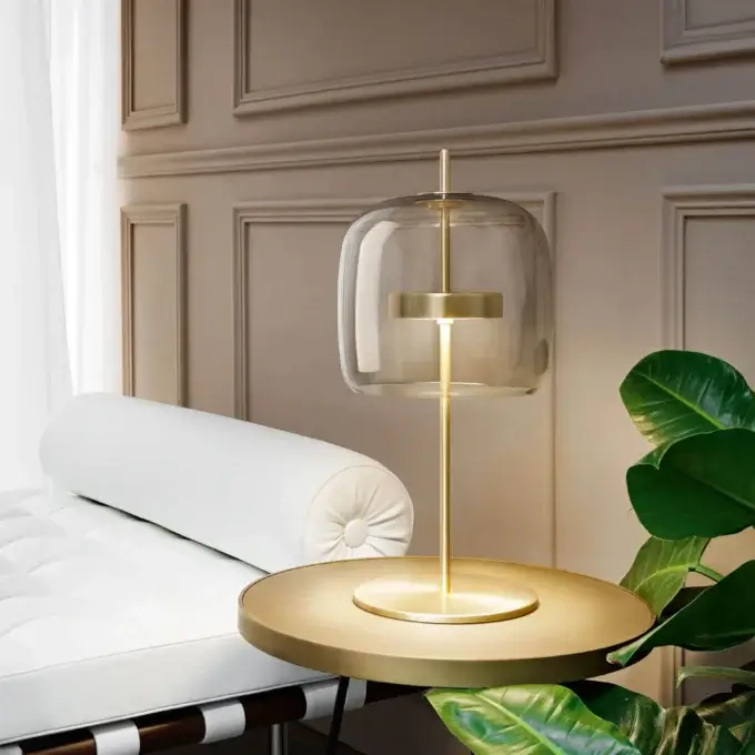 Vistosi table lamp with brass base and amber glass shade, beside white chaise lounge and green plant on dark surface.