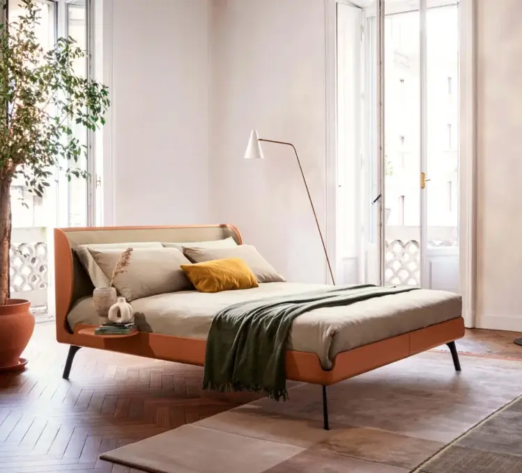 Bolzan Letti gabri bed, beige fabric headboard, rust frame, brass legs, gray linen bedding, green throw, arched floor lamp, hardwood floor.