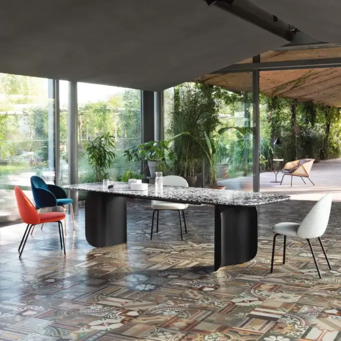 Dining table with dark grey and white mosaic top, surrounded by orange and white chairs, in a room with patterned terracotta flooring and glass walls.