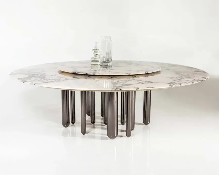 Carrara marble dining table with dark gray metal legs and brass rotating base, featuring gray veining and clear glass vases.