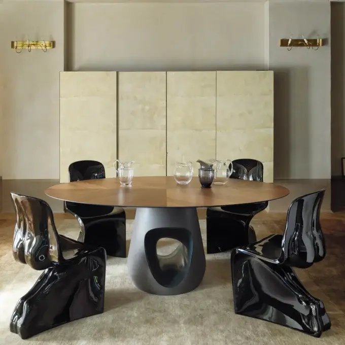 Round dining table with light wood grain, six black sculptural chairs, brass sconces, beige tile wall panel, and light beige rug.