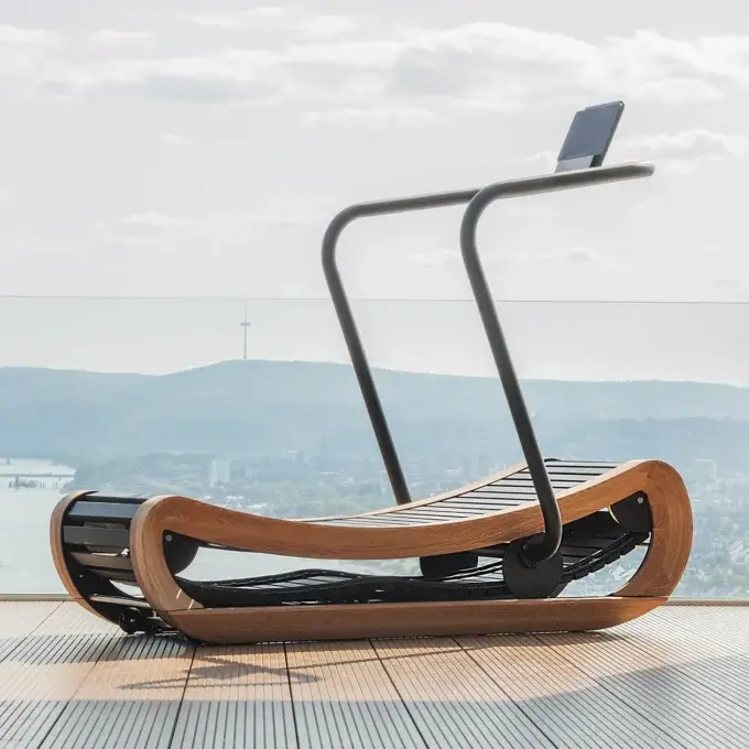 Curved treadmill with light wood frame, black metal base, and integrated screen, positioned on a deck overlooking a city landscape.