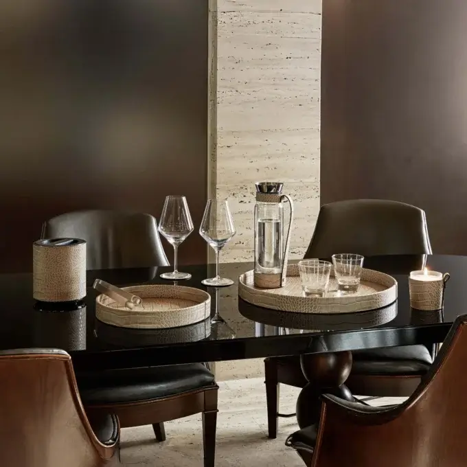 Black lacquered round dining table with crocodile-texture barware, dark leather armchairs, beige travertine floor, and cream wall paneling.